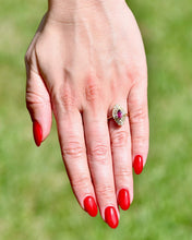 Load image into Gallery viewer, Hand with red nail polish wearing a ring with a ruby surrounded by diamonds against a blurred green background