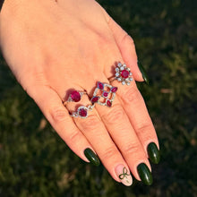 Load image into Gallery viewer, Hand wearing multiple ruby and diamond rings and green nail polish against a blurred natural background