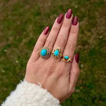 Load image into Gallery viewer, Hand wearing three gold rings with turquoise stones against a grassy background.
