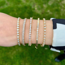 Load image into Gallery viewer, Wrist wearing multiple yellow gold diamond tennis bracelets with a green outdoor background