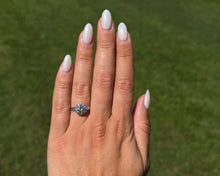 Load image into Gallery viewer, A model wearing a signed Verragio 14K white gold engagement ring featuring a round brilliant-cut center diamond with a halo setting and split shank, shown outdoors on a hand against a soft, blurred green lawn background.
