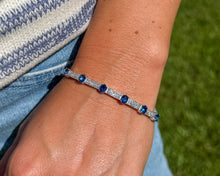 Load image into Gallery viewer, A model wearing an 18K white gold flexible cuff bracelet with alternating oval blue sapphires and pavé diamond links, set against a knitted sweater and blue jeans.
