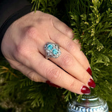 Load image into Gallery viewer, Hand wearing an antique platinum blue zircon and diamond ring against a green foliage background.
