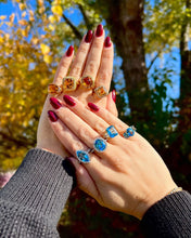 Load image into Gallery viewer, A hand wearing multiple citrine rings and the other hand wearing multiple blue topaz rings against a blurred natural background.
