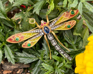 Vintage 18k yellow gold and sterling silver dragonfly brooch set with blue sapphire, diamonds, emeralds, and rubies pictured on a leaf.