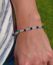 Load and play video in Gallery viewer, Video of a model wearing an 18K white gold flexible cuff bracelet with alternating oval blue sapphires and pavé diamond links, set against a knitted sweater, blue jeans, and an outdoor green background.
